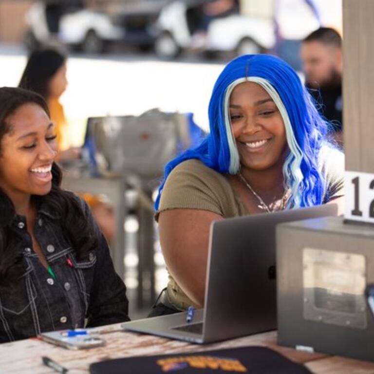 A woman with blue hair and a woman with black hair are sitting next to each other and laughing.