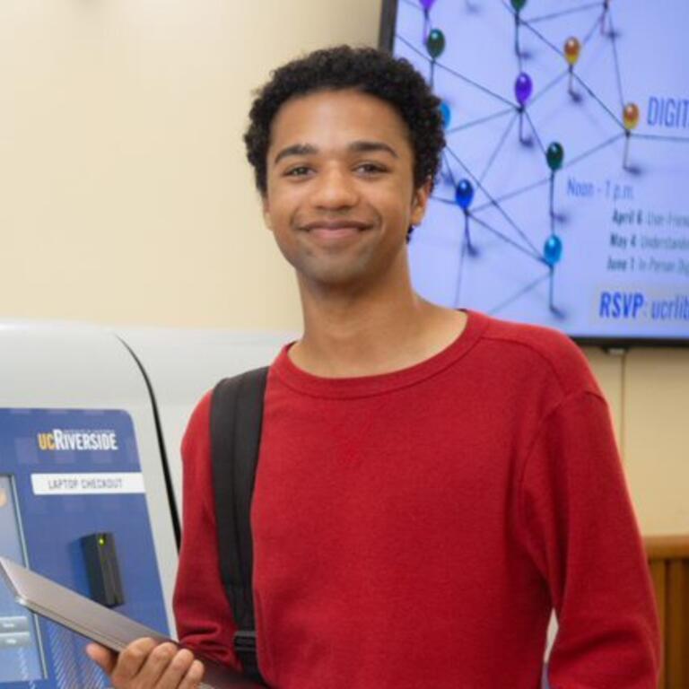A student in a red shirt is smiling while holding a laptop.