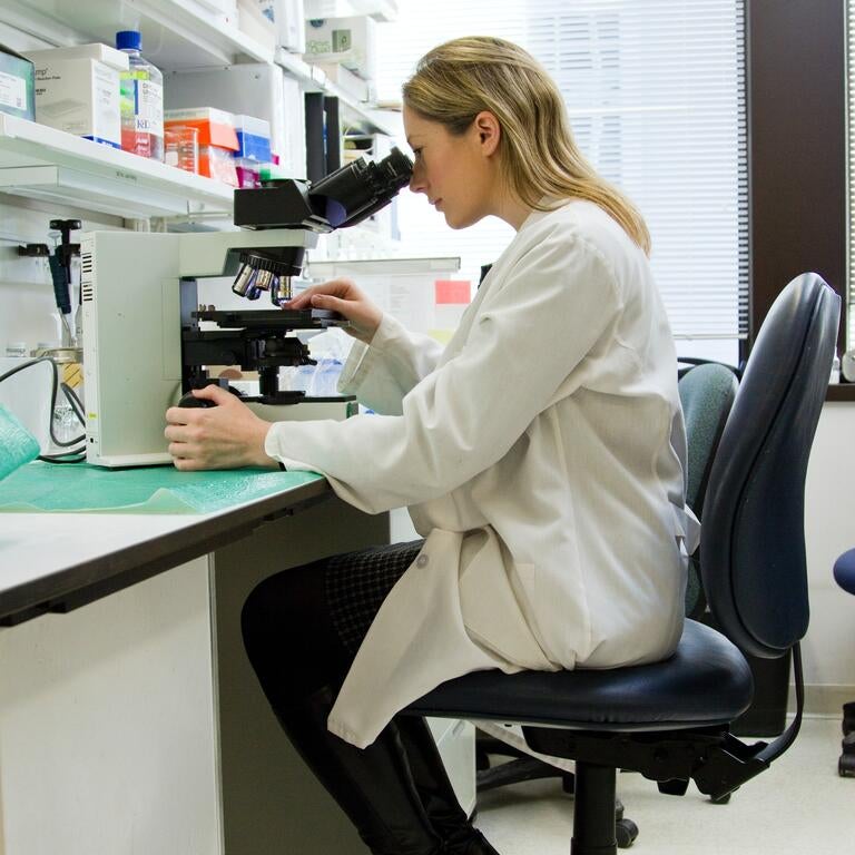 A blonde woman in a lab coat is looking into a microscope.