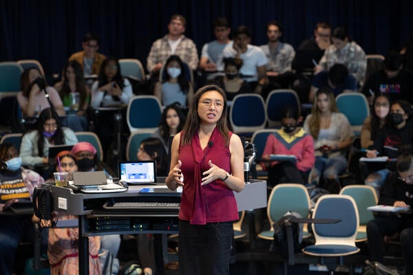 A faculty member in a red shirt and glasses lecturing to a room of students.