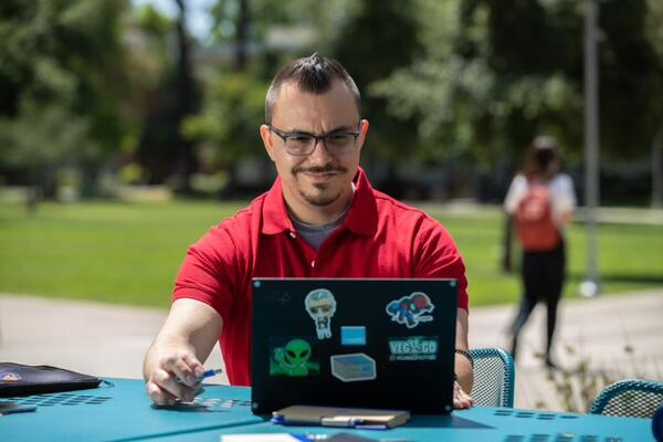 A staff member in a red shirt sits behind his laptop and smiles