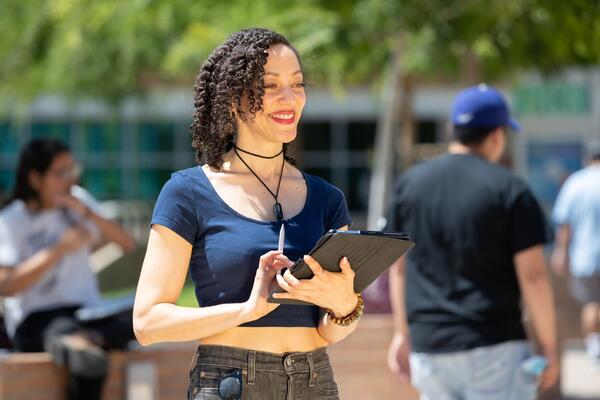 A woman in a blue shirt is holding a tablet and pen while standing and smiling.