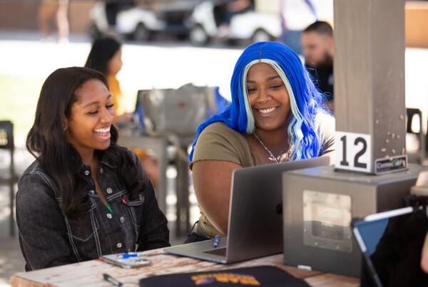 A woman with blue hair and a woman with black hair are sitting next to each other and laughing.