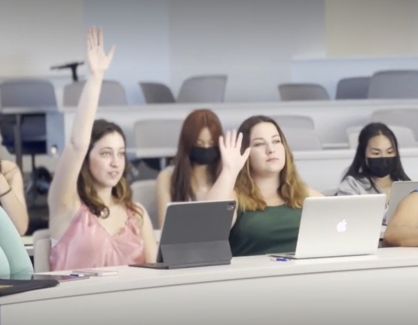 a student in a pink shirt and her classmate in a green tank top raising their hands in class