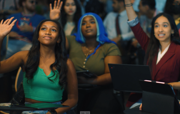 A group of students raising their hands in class. One student is wearing green. Another student with blue hair is wearing an olive t-shirt. Another student is wearing a red blazer.