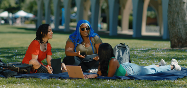 Three students sitting on the grass in front of Rivera Library with a computer in front of them.