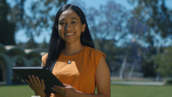 A woman in a yellow dress and long dark hair is smiling while holding a tablet.