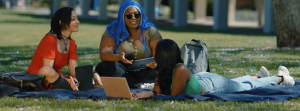 Three students sitting on the grass in front of Rivera Library with a computer in front of them.