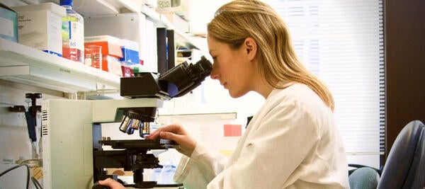 A blond woman wearing a lab coat looking into a microscope while working in a lab