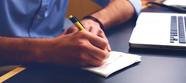 A man's hand takes note on paper looking at laptop.