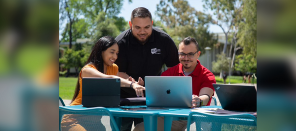 A worker in a black ITS Staff Shirt assisting a man and a woman with their laptops